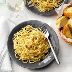 two black plates with spaghetti and bread on them next to wine glasses, napkins and utensils