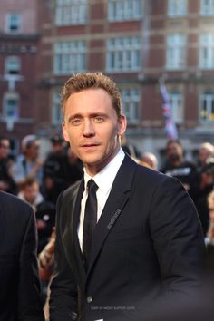 a man in a suit and tie standing on a red carpet with people behind him