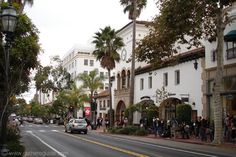 people are standing on the sidewalk in front of buildings and palm trees, with one car driving down the street