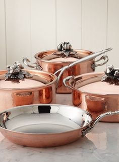 four copper pots and pans sitting on a marble counter top with silverware in the middle