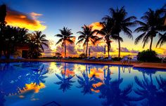 a pool with palm trees and the sun setting in the background, reflecting on the water