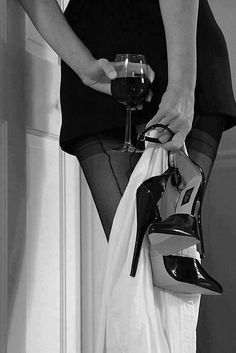 a woman holding a wine glass in her right hand while standing next to a pair of high heeled shoes