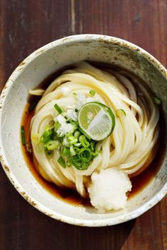 a white bowl filled with noodles and garnished with green onions, scallions and sliced limes