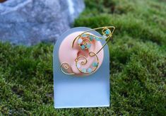 a pink and gold brooch sitting on top of a green grass covered ground next to a rock