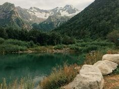 a man standing on the side of a mountain next to a lake with mountains in the background