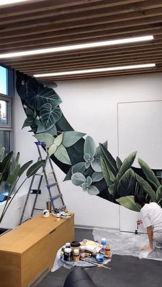 a man painting a mural on the wall in an office space with plants and other things