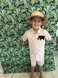 a young boy wearing a hat and overalls standing in front of a wall with green leaves