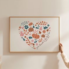 a woman is holding up a framed art print with flowers and pumpkins in the shape of a heart