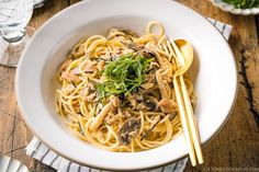 a white bowl filled with pasta and mushrooms on top of a wooden table next to chopsticks