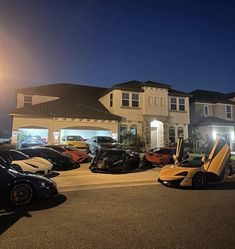 several exotic sports cars are parked in front of a luxury house at night with the lights on