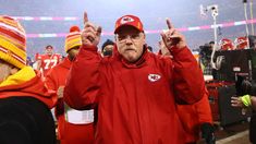 a man in a red jacket and hat holding up two fingers at a football game