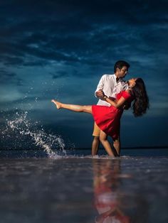 a man and woman dancing on the beach at night with water splashing around them