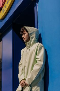 a man in a white raincoat leaning against a blue wall