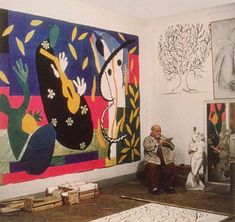 an older man sitting in front of paintings on the wall and another person standing next to him