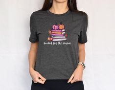 Booked for the Season T-shirt, perfect for book lovers and autumn enthusiasts alike! This Halloween tee features a vibrant graphic of a colorful stack of books, accented with pumpkins to capture the warm, cozy vibes of fall. Whether you're curling up with a good read or enjoying a pumpkin spice latte, this soft, comfortable fall shirt will be your go-to for the season. Ideal for library visits, casual outings, or cozy nights in, it's a must-have for anyone who loves combining their passion for r Fall Bookish Tops With Graphic Print, Fall Graphic Print Bookish Tops, Fall Book Shirt, Bookish Fall T-shirt With Letter Print, Fall Bookish T-shirt With Letter Print, Relaxed Fit Pre-shrunk Bookish T-shirt, Bookish Cotton T-shirt With Slogan, Bookish Cotton T-shirt With Funny Print, Book Lover Shirts Inspire Uplift ⭐