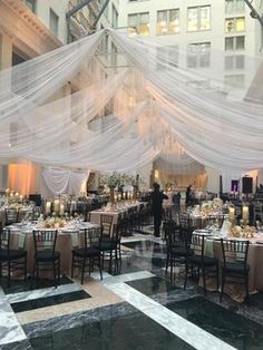 an indoor banquet hall with tables and chairs covered in white sheer cloths, surrounded by tall buildings