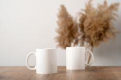 two white mugs sitting on top of a wooden table next to a dried plant