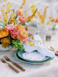 an arrangement of flowers and napkins on a white table cloth with gold cutlery