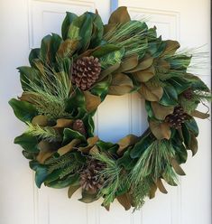 a green wreath with pine cones and evergreens hanging on a white front door,