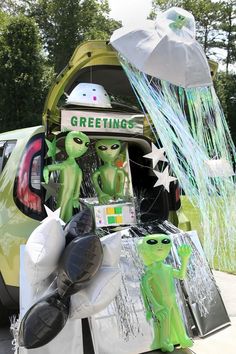 an open trunk with green alien figures and balloons attached to it's doors, in front of a parked car