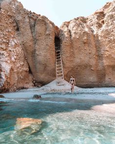 a woman standing on the beach next to a cliff with a ladder going up it's side