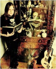 a man holding a book standing in front of a dresser with guitars and skulls on it