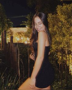 a woman in a black dress posing for the camera at night with trees and bushes behind her