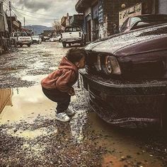 a young child standing next to a parked car on a wet street with cars in the background