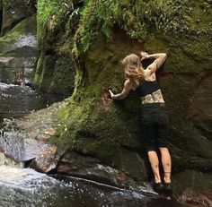 a woman standing on top of a mossy rock next to a river in the forest