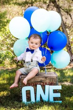 a baby sitting on top of a wooden crate holding blue and white balloons in the air