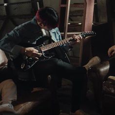 a man sitting in a chair playing an electric guitar with another person standing next to him