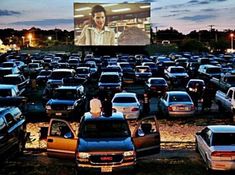 a large group of cars parked in a parking lot with a movie screen behind them