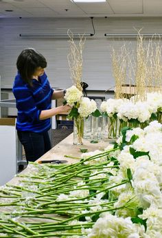 a woman arranging white flowers in vases on a table with another person looking at them