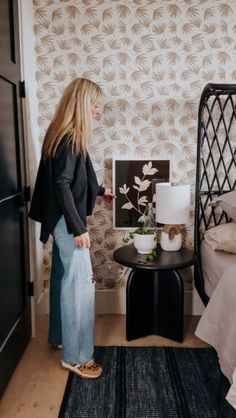 a woman standing next to a bed in a room with a black table and white wallpaper