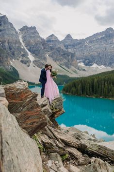 a bride and groom standing on the edge of a cliff overlooking a mountain lake