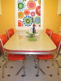 a dining room table with red chairs and a vase on the top, in front of a yellow wall