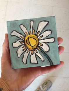 a hand holding up a painted tile with a yellow and white flower in the center