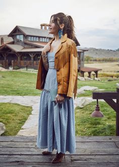 a woman standing on a wooden deck wearing a blue dress and brown jacket with fringes