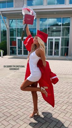 a woman in a red and white graduation outfit holding up a book while standing on a brick walkway