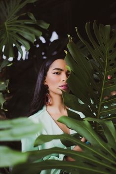 a woman with long black hair is standing in front of some plants and looking at the camera