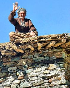 an old man sitting on top of a stone building waving at the camera with his hand