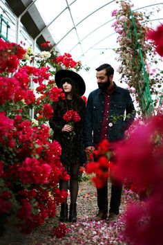 a man and woman walking through a greenhouse with red flowers in the foreground, looking at each other