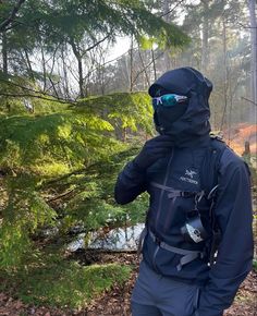 a man in a black jacket and goggles standing next to a forest with trees