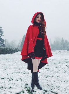 a woman standing in the snow wearing a red coat and black dress with hood on