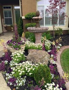 a garden with flowers and rocks in front of a house that has a water fountain