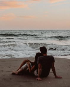 two people are sitting on the beach watching the waves come in to shore as the sun sets