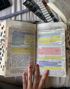 a person's hand holding an open book on top of a bed next to other books