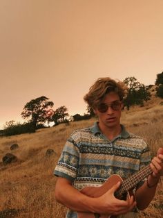 a man standing in a field holding a guitar and looking at the camera with an orange sky behind him