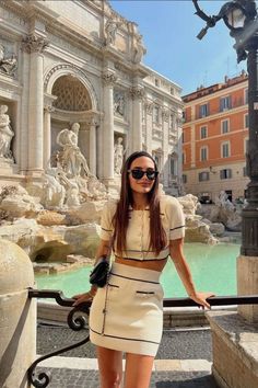 a woman standing in front of a fountain with her hands on her hips and wearing sunglasses