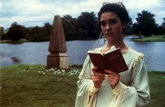 a woman dressed in white holding a book and looking at the camera while standing next to a lake